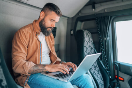 Captured inside his truck, a truck driver is seen seated on the bed while focused on his laptop, managing tasks in the limited space of his vehicle